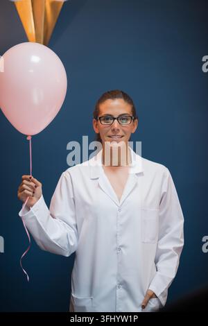Junge medizinische Technikerin in einem weißen Laborkittel hält einen rosa Ballon vor einem blauen Hintergrund und verbindet professionelle klinische Präsenz mit einem leichten Stockfoto