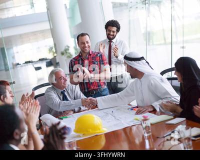Nahost-Profis aus Unternehmen und Startups schütteln sich die Hände über Bauplänen und einem Sicherheitshut und applaudieren einer erfolgreichen Vereinbarung. Das diverse Team ref Stockfoto