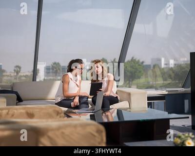 Zwei professionelle Frauen aus einem Startup im Nahen Osten arbeiten in einer modernen Bürolounge zusammen, überprüfen Dokumente auf einem Tablet und Laptop und diskutieren Stockfoto