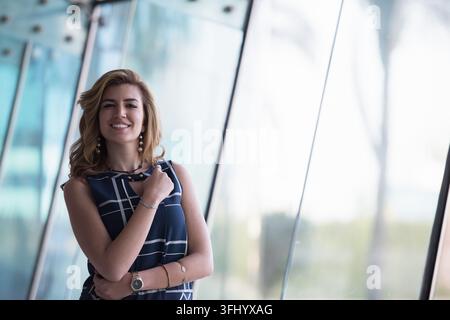 Eine selbstbewusste junge Geschäftsfrau lächelt, während sie neben großen Glasfenstern in einem modernen Büro steht, und trägt ein gemustertes ärmelloses Kleid und Schmuck; nat Stockfoto