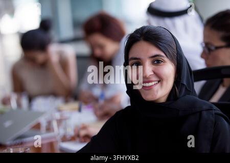 Lächelnde Geschäftsfrau mit Hijab nimmt selbstbewusst an einer vielfältigen Teambesprechung teil, umgeben von Kollegen, Laptops und Notizen. Kollaborativ w Stockfoto