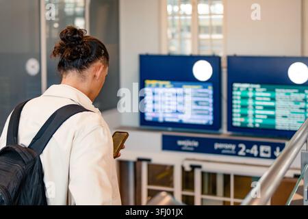 Geschäftsreisender überprüft Zuginformationen auf einer Bahnhofsanzeige, während er sein Smartphone nutzt. Konzept von Geschäftsreisen, Reiseplanung und Digitalität Stockfoto