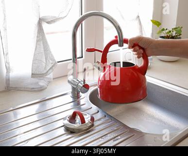 Frauenhände füllen einen roten Wasserkocher mit Leitungswasser im Küchenwaschbecken. Stockfoto