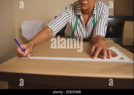 Eine Frau misst und markiert ein Papierschnittmuster auf braunem Stoff mit einem Lineal und Maßband, um ein Kleidungsstück für die Schneiderei oder Kleiderherstellung vorzubereiten Stockfoto