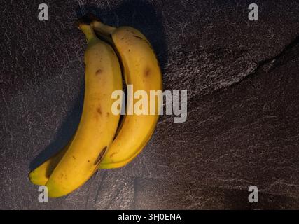 Eine Nahaufnahme, hochwinklig aufgenommen, zeigt einen kleinen Bunch frischer, reifer gelber Bananen, die diagonal über einen dunklen, rauen und strukturierten Stein ruhen, s Stockfoto