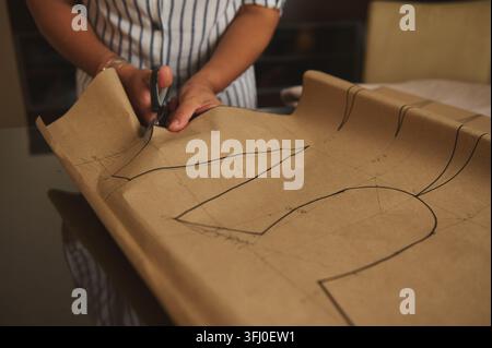 Frau schneidet ein großes braunes Papierschnittmuster mit einer Schere auf einem Tisch und zeigt das Anfertigen von Schnittmustern, Nähen, Schneidern und DIY-Handwerk in einem Heimbüro Stockfoto