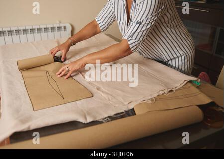 Frau legt ein Papiermuster aus und schneidet leichten Stoff auf einem Tisch, veranschaulicht Schneiderei, Maßschneiderei und Heimnähhandwerk mit Maßband an Stockfoto