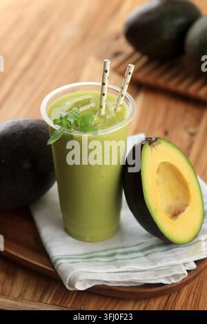 Frischer Avocado-Smoothie in einem klaren Plastikbecher. Grünes Getränk mit Minzblättern und gestreiften Strohhalmen garniert. Halbierte Avocado neben dem Becher zeigt das cremige Gelb Stockfoto