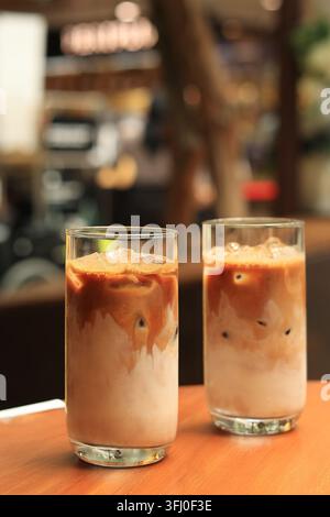 Zwei Eiskaffee-Getränke Kopi Susu Gula Aren auf einem Holztisch. Braune Milch wirbelt um Eiswürfel in klarem Glas. Weicher Bokeh-Hintergrund deutet auf ein Café hin Stockfoto