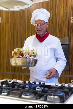 Ein lächelnder nahöstlicher Koch in weißer Uniform und Toque wirft Gemüse in einer Pfanne über einem Gasherd, während er das Iftar-Dinner für eine Familienfeier zubereitet Stockfoto