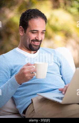 Moderner arabischer Mann aus dem Nahen Osten sitzt draußen mit Laptop und Kaffee, lächelt und entspannt, während er von zu Hause aus auf einer sonnigen Terrasse, Casua, arbeitet Stockfoto
