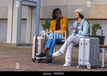 Verschiedene Freunde sitzen auf einer Holzbank am plaza und unterhalten sich mit Koffern und Rucksäcken. Reisende, Gepäck, Outdoor, Urban, Innenhof, Sitzplätze, plante Stockfoto