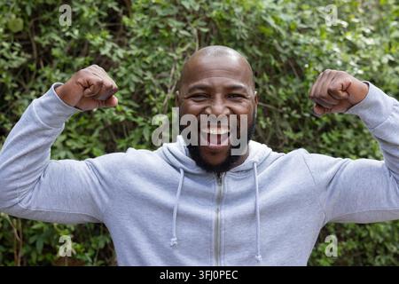 Afroamerikaner lächelt und beugt sich, während er in einer Hinterhofhecke mit grauem Hoodie steht. Im Freien, Grün, Garten, Sträucher, Tageslicht, lebendig, Stockfoto