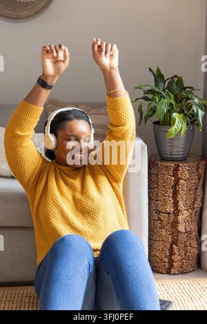 Afroamerikanerin sitzt auf einem Teppich im Wohnzimmer, trägt senfgelbe Strick-Kopfhörer, hebt die Arme. Person, Musik, gemütlich, beige, Stockfoto