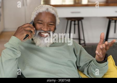 Ranghoher Afroamerikaner lächelt, spricht auf dem Smartphone, während er auf dem Sofa mit gelbem Kissen sitzt. Ältere Menschen, Telefon, Couch, Sonnenlicht, offener Plan, Küche, Stockfoto