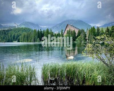 Panoramatische Ansicht von Touristen, die rote Boote auf dem Štrbské Pleso-See in der Hochtatra in der Slowakei rudern, mit Hotel Patria und nebligen Bergen im Hintergrund. Stockfoto