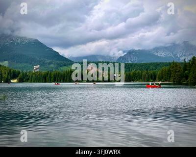 Panoramatische Ansicht von Touristen, die rote Boote auf dem Štrbské Pleso-See in der Hochtatra in der Slowakei rudern, mit Hotel Patria und nebligen Bergen im Hintergrund. Stockfoto