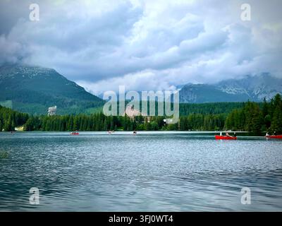 Panoramatische Ansicht von Touristen, die rote Boote auf dem Štrbské Pleso-See in der Hochtatra in der Slowakei rudern, mit Hotel Patria und nebligen Bergen im Hintergrund. Stockfoto