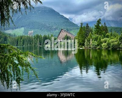Panoramatische Ansicht von Touristen, die rote Boote auf dem Štrbské Pleso-See in der Hochtatra in der Slowakei rudern, mit Hotel Patria und nebligen Bergen im Hintergrund. Stockfoto