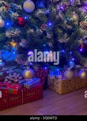 Weihnachtsgeschenke, die auf einem Holzboden unter einer mit farbenfrohen Kugeln und lebhaften LED-Lichtern geschmückten Kiefern platziert sind, spiegeln eine gemütliche Winterstimmung während Stockfoto