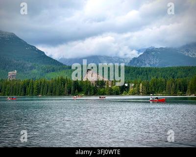 Panoramatische Ansicht von Touristen, die rote Boote auf dem Štrbské Pleso-See in der Hochtatra in der Slowakei rudern, mit Hotel Patria und nebligen Bergen im Hintergrund. Stockfoto