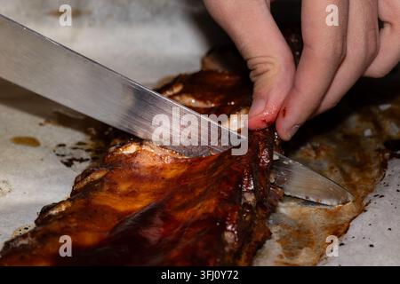 Nahaufnahme von gegrillten Barbecue-Rippchen, die während der Essenszubereitung mit einem Küchenmesser geschnitten werden, und präsentieren gegartes Fleisch, knusprige Textur und Hausmannskost. Stockfoto