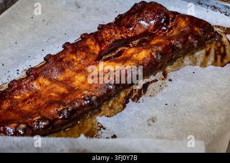 Nahaufnahme von glasierten Schweinerippchen, die auf Backpapier in einer Ofenschale gebacken wurden, und zeigen saftiges Barbecue-Fleisch, das zum Verzehr bereit ist. Stockfoto