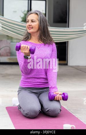 Frau mittleren Alters kniend auf der Terrassenmatte unter der Hängematte, lila Kurzhanteln haltend, lila Top. Outdoor, Fitness, Gewichte, Terrasse, Beton, gar Stockfoto