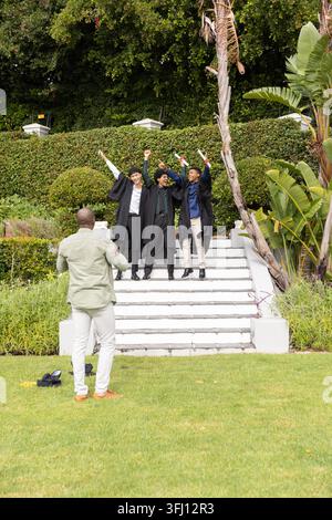 Männliche Absolventen in schwarzen Gewändern mit Diplomen auf den Stufen, afroamerikanischer Mann filmt mit Telefon. Abschluss, im Freien, Garten, Treppe, Diplom, p Stockfoto