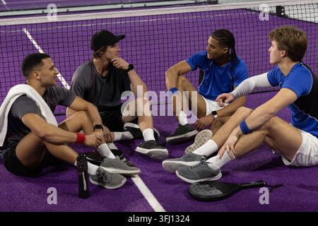 Verschiedene 4 männliche Athleten sitzen im Netz auf dem violetten Platz in Sportbekleidung mit Paddel, plaudern. Pickleball, Teamkollegen, Kameradschaft, Gelegenheitsgebrauch, Konversation, Stockfoto