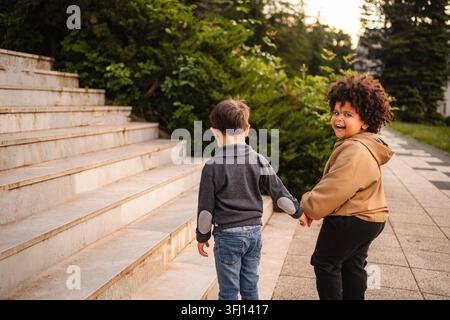 Zwei verschiedene Kinderfreunde halten Hände, gehen auf Treppen in einem Park und lächeln Stockfoto