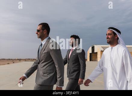 Drei professionelle Männer, zwei in Anzügen und einer in traditionellem Emirat-Kandura, gehen zusammen auf einem Flughafenparkplatz, vermitteln Teamarbeit, Selbstvertrauen, Korpora Stockfoto