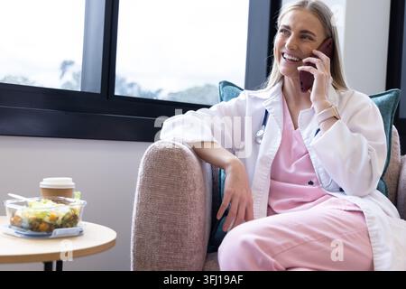 Ärztin sitzt am Fenster in der Personallounge, Laborkittel mit Stethoskop, Smartphone in der Hand. Medizin, Klinik, Ruhe, Sessel, Tageslicht, tragbar Stockfoto