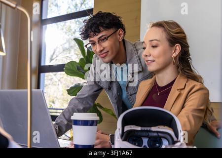 Verschiedene Kollegen arbeiten am Laptop am Schreibtisch in der Nähe des Fensters mit VR-Headset und Kaffeetasse zusammen. Teamwork, Büro, Coworking, Zusammenarbeit, modern, Dayl Stockfoto