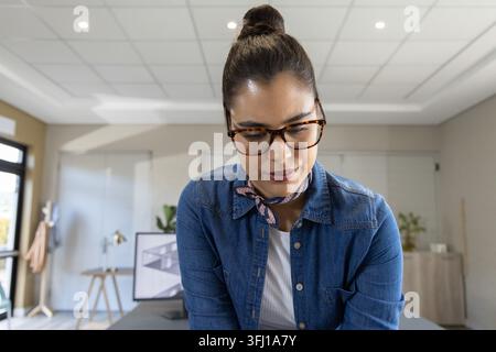 Erwachsene Frau lehnt sich über den Schreibtisch in einem modernen Studio, trägt Schildpatt-Brille und Denim-Hemd. Professionell, Architekt, Designer, Fokus, Konzentration Stockfoto