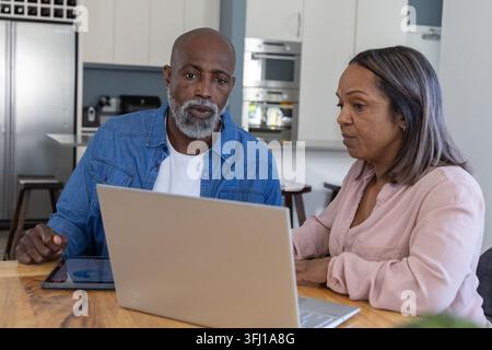 Älteres afroamerikanisches Paar mit silbernem Laptop und Tablet am hölzernen Esstisch in der Küche. Reife, Duo, Heim, modern, Interieur, Geräte, Fokus Stockfoto