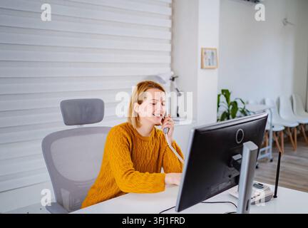 Junge Frau lächelt, spricht am Bürotelefon, arbeitet Stockfoto