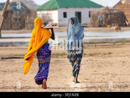 Zwei Frauen tragen Tabletts mit Muschelhalsketten auf ihren Schultern, während sie durch ein Küstendorf im Nördlichen Roten Meer, Dahlak, Eritrea, gehen Stockfoto