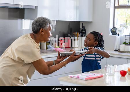 Afroamerikaner Seniorin und Kind Mädchen, die Metallbacktablett in der Küche, Denimschürze. Zusammenarbeit, generationenübergreifend, kochen, Familie Stockfoto
