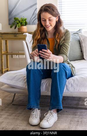 Frau, die zu Hause auf Futon sitzt, lächelt und auf ein marineblaues Smartphone blickt, in Jeans mit Olivhemd. Erwachsene, Innenraum, gemütlich, Tageslicht, Pflanze, abstrakt, min Stockfoto