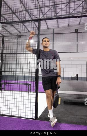 Erwachsener Mann mit grauem Hemd, gestützt auf schwarzem Mesh, mit Paddel, stehend auf lila Court. Schläger, Käfig, Einrichtung, Beleuchtung, Fitnessstudio, Freizeit-, Sport Stockfoto