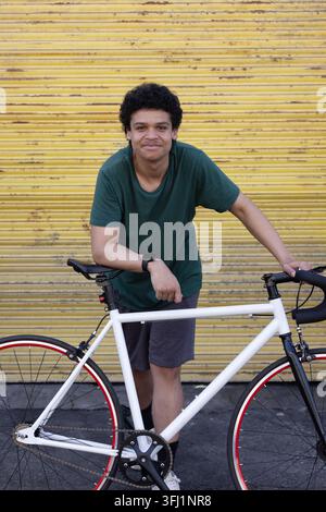 Afroamerikanischer Mann lehnt sich an ein weißes Rennrad vor einem gelben Fensterladen, trägt eine Smartwatch. Fahrrad, urban, Ladenfront, Pflaster Stockfoto