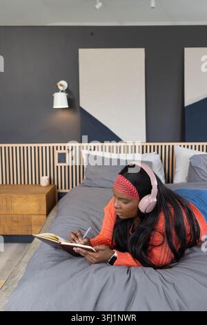 Erwachsene afroamerikanische Frau liegt auf dem Bett und schreibt im Schlafzimmer mit einem Notizbuch, trägt rosa Headset Stockfoto