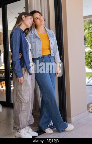 Verschiedene weibliche Partner stehen an der Glasschiebetür und lehnen sich in Jeansjacken zusammen. Frauen, Sonnenlicht, Tür, Pool, Jeans, Home, Compani Stockfoto
