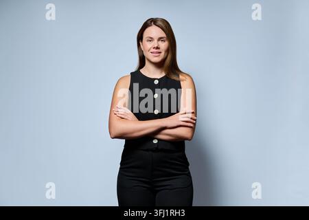 Porträt einer selbstbewussten jungen Frau in einem schwarzen ärmellosen Oberteil, die mit verschränkten Armen vor einem Studiohintergrund steht. Konzept moderner Führung, profes Stockfoto