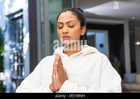Mittelerwachsene Indianerin, die zu Hause in der Nähe der Glastür steht, mit weißem Hoodie, die Handflächen drücken auf die Brust. Meditation, Achtsamkeit, Gelassenheit, Atmen, mudr Stockfoto