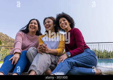 Verschiedene Freundinnen lachten und lehnten sich auf der Poolterrasse mit Pullover und Jeans und Smartwatch. Trio, Outdoor, Freundschaft, offen, sonnig, lächelt, Stockfoto