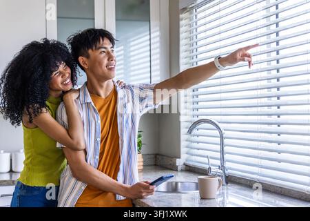 Afroamerikanerin und asiatischer Mann, der an der Marmortheke steht und das Telefon auf Jalousien zeigt. Küche, Paar, modern, Tageslicht, Smartphone, zu erhalten Stockfoto