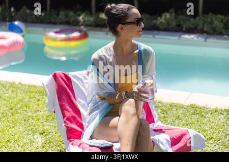 Frau mittleren Alters, die auf einem mit Handtüchern überzogenen Liegestuhl am heimischen Pool sitzt, in gelben Badeanzügen mit einem Glas. Garten, sonnendurchflutete, Entspannung, draußen, Sonnenbrille, Stockfoto
