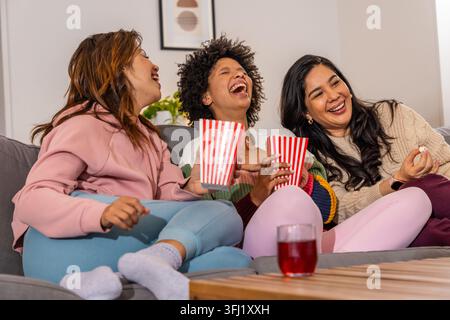 Verschiedene Freundinnen lachen zusammen und teilen gestreifte Popcorn-Wannen auf grauem Sofa im Wohnzimmer. Vielfältig, Freundinnen, Lachen, Snacks, Couch, Startseite Stockfoto